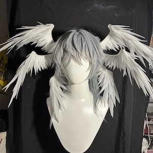 May include: A white mannequin head wearing a layered, light gray wig styled with bangs. Attached to the wig are four white, feathered-style wings extending outward, with two larger wings positioned at the top and two smaller wings at the bottom.