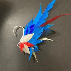 May include: A vibrant ear cuff featuring a cluster of colorful feathers. The design incorporates bright blue, red, white, and black feathers, attached to a silver-toned metal ear cuff. The feathers are arranged to create a striking, artistic accessory.