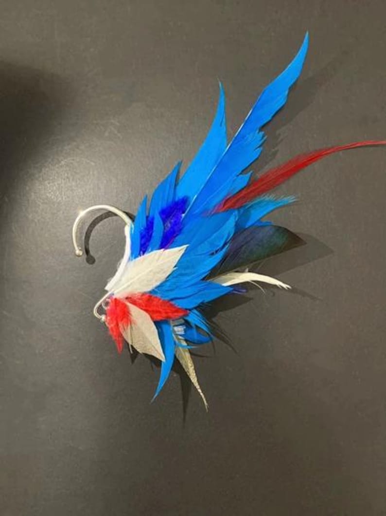 May include: A vibrant ear cuff featuring a cluster of colorful feathers. The design incorporates bright blue, red, white, and black feathers, attached to a silver-toned metal ear cuff. The feathers are arranged to create a striking, artistic accessory.