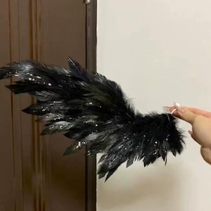 May include: A black feather wing with glitter accents. The wing is made of multiple black feathers and is held up against a white wall and a brown door. The feathers are arranged to resemble a wing.