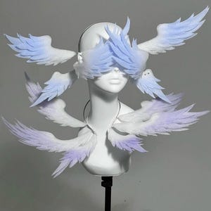 May include: A white mannequin head is adorned with a fantasy-themed costume. The costume features multiple pairs of feathery wings in shades of white and light blue. The wings are arranged around the eyes and neck, creating a unique, ethereal look.