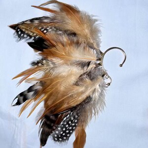 May include: An ear cuff featuring a cluster of brown, tan, black, and white feathers. The feathers are attached to a silver-colored metal ear cuff with a curved design. The feathers have a variety of patterns and textures.