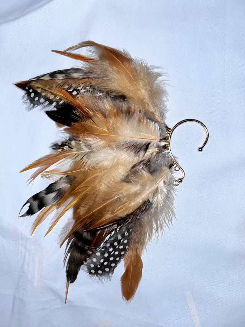 May include: An ear cuff featuring a cluster of brown, tan, black, and white feathers. The feathers are attached to a silver-colored metal ear cuff with a curved design. The feathers have a variety of patterns and textures.