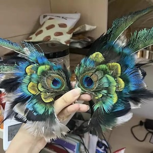 Puede incluir: Un par de m&aacute;scaras para los ojos de plumas de pavo real. Las m&aacute;scaras presentan una combinaci&oacute;n de plumas azules, verdes, amarillas y negras, con patrones intrincados. Las m&aacute;scaras son sostenidas por una mano, mostrando la artesan&iacute;a y el dise&ntilde;o detallados.