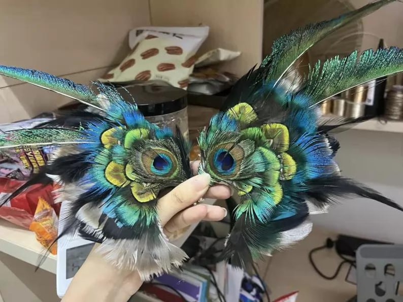 Puede incluir: Un par de m&aacute;scaras para los ojos de plumas de pavo real. Las m&aacute;scaras presentan una combinaci&oacute;n de plumas azules, verdes, amarillas y negras, con patrones intrincados. Las m&aacute;scaras son sostenidas por una mano, mostrando la artesan&iacute;a y el dise&ntilde;o detallados.