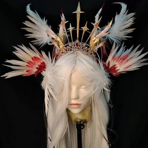 May include: A headpiece featuring a gold-toned metal crown adorned with small red gems, topped with multiple gold star-shaped spikes. The design includes large, layered white feathers with red and blue tips extending outward from the sides.