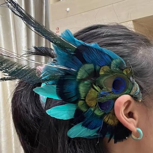Puede incluir: Un tocado vibrante con plumas de pavo real en tonos turquesa, azul y verde. Las plumas est&aacute;n dispuestas en forma de abanico, con algunas que se extienden hacia arriba. El tocado se lleva en el lateral de la cabeza, cerca de la oreja.