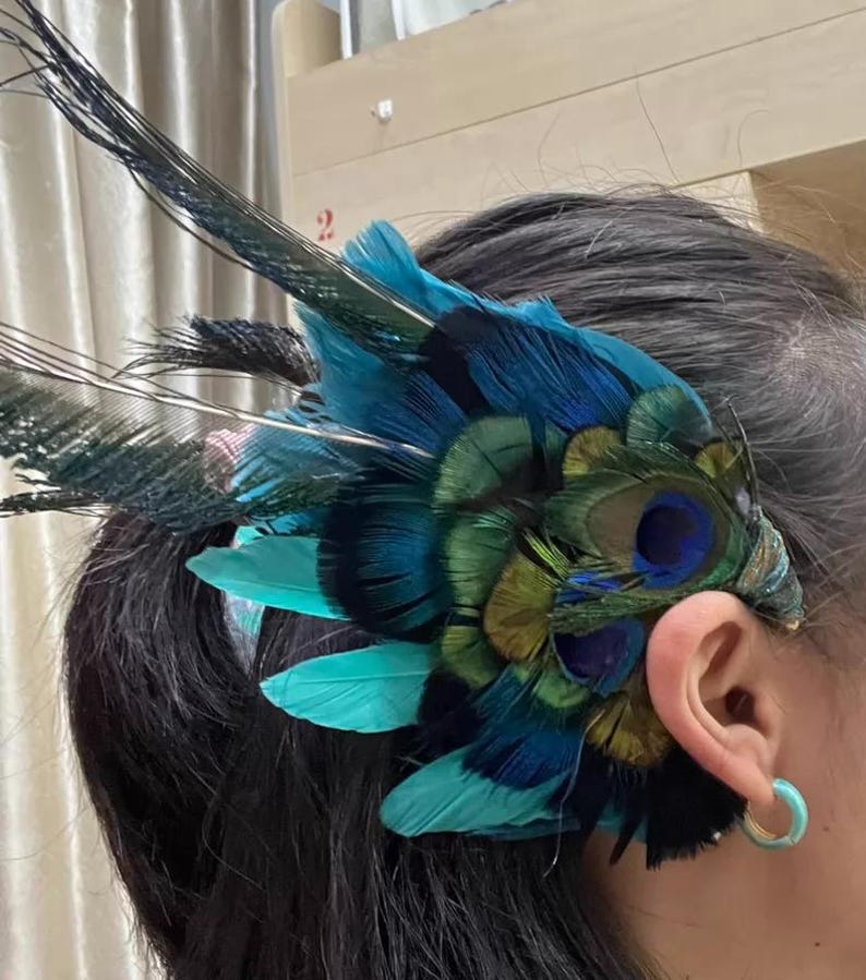 Puede incluir: Un tocado vibrante con plumas de pavo real en tonos turquesa, azul y verde. Las plumas est&aacute;n dispuestas en forma de abanico, con algunas que se extienden hacia arriba. El tocado se lleva en el lateral de la cabeza, cerca de la oreja.