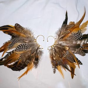 May include: A pair of ear cuffs featuring a wing design made of brown, tan, and black feathers. The feathers are attached to a gold-colored metal ear cuff. The feathers have white polka dots.
