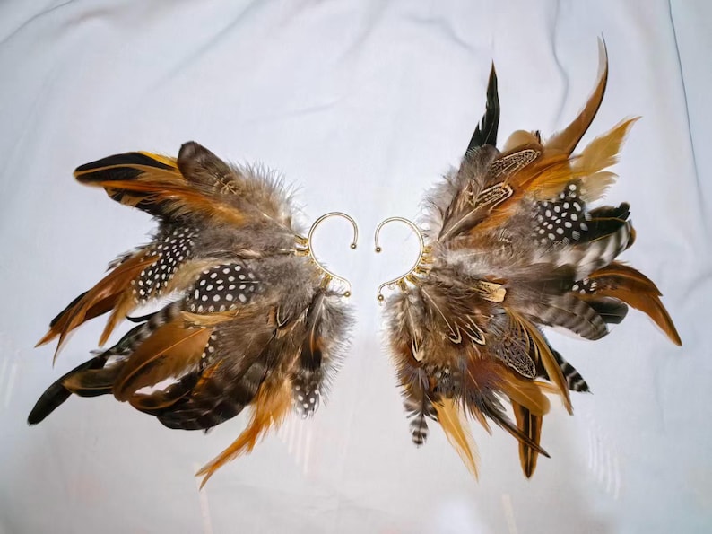 May include: A pair of ear cuffs featuring a wing design made of brown, tan, and black feathers. The feathers are attached to a gold-colored metal ear cuff. The feathers have white polka dots.