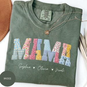 May include: Moss green t-shirt with the word "MAMA" in patchwork letters. The letters are decorated with floral and gingham patterns. Below the word "MAMA" are the names Sophia, Olivia, and Noah. A gold necklace is draped on the shirt.
