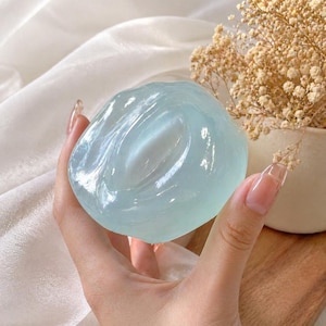 May include: A translucent, light blue, oval-shaped soap bar. The soap has a smooth, glossy surface and is held in a hand. The background includes a white fabric and a small vase with dried flowers.