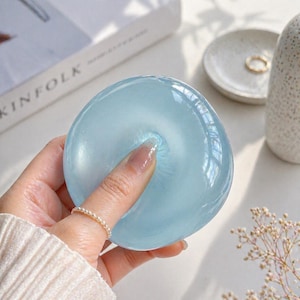 May include: A light blue, round, glass object is held by a hand. The object is smooth and translucent, with a finger pressing into its center. A pearl ring adorns the finger. Other items in the background include a book and a small dish.