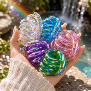 May include: A collection of five iridescent, coiled plastic springs in clear, blue, purple, pink, and green colors. Each spring features a smooth, segmented, and translucent design that reflects light, creating a shimmering effect across the surfaces.