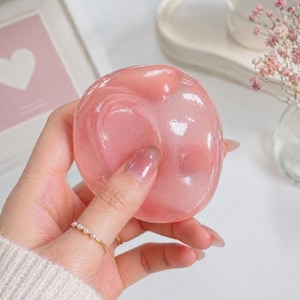 May include: A translucent, pink, heart-shaped object is held in a hand. The object has a glossy appearance. A gold ring with small pearls is on the ring finger. The background includes a framed picture and a vase with pink flowers.