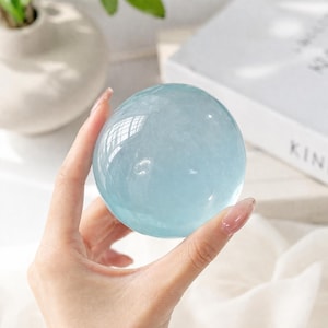 May include: A clear, light blue crystal sphere, held in a hand. The sphere reflects light, creating a shimmering effect. The crystal ball is a popular item for home decor and spiritual practices.