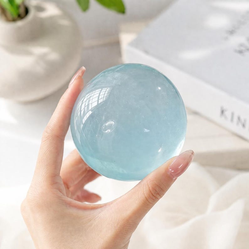 May include: A clear, light blue crystal sphere, held in a hand. The sphere reflects light, creating a shimmering effect. The crystal ball is a popular item for home decor and spiritual practices.