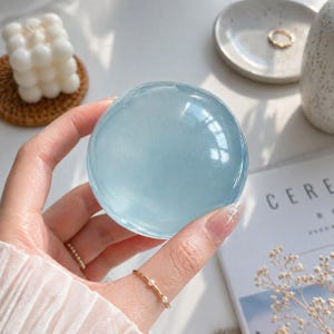 May include: A translucent, light blue sphere is held in a hand. The sphere reflects light, creating highlights. A gold ring with pearls and a gold ring are visible on the fingers. A small white candle and a ceramic dish are in the background.