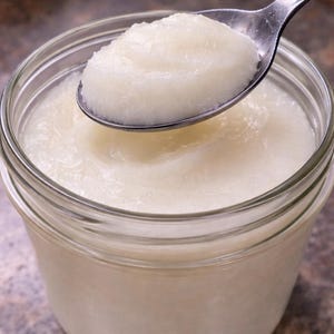 May include: A clear glass jar filled with a thick, white substance. A silver spoon scoops a portion of the white substance from the jar. The jar is sitting on a brown surface.