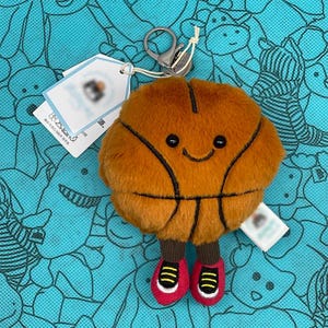 Peut inclure: Un porte-clés en forme de ballon de basket en peluche avec un visage souriant, des lignes noires et des chaussures rouges à rayures jaunes. Le porte-clés a un fermoir argenté et une étiquette attachée. Le fond est un motif bleu clair.