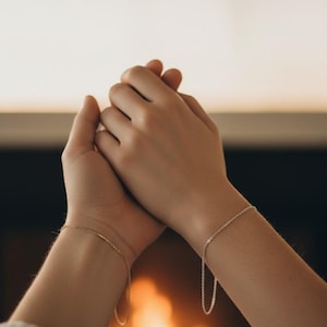 May include: Two wrists with delicate chain bracelets, one gold and one silver, are clasped together. The gold bracelet features a small bar, while the silver one has a simple chain design. The background is softly blurred with warm tones.