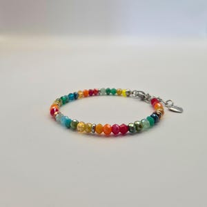 May include: A colorful beaded bracelet featuring a spectrum of colors, including red, orange, yellow, green, and blue. The bracelet has a silver-toned clasp and a small heart charm. The beads are small and faceted, creating a sparkling effect.