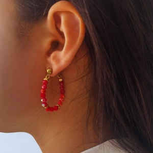 Puede incluir: Pendientes de aro rojos con cuentas y detalles dorados. Los pendientes tienen forma ovalada y presentan pequeñas cuentas rojas facetadas. Los pendientes son una joya.