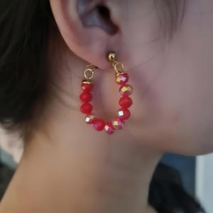 Può includere: Orecchini a cerchio con perline rosse sfaccettate. Gli orecchini hanno un perno in metallo color oro e un piccolo cerchio color oro che collega il perno al cerchio di perline. Le perline sono di un rosso vivace.