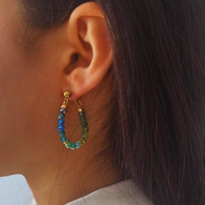 May include: Gold-tone hoop earrings with a gradient of blue and green beads. The earrings are attached to a small gold stud. The beads are small and round, creating a delicate and colorful accessory.