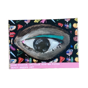 May include: Hand-painted, upcycled sew-on patch featuring a large eye design. The eye is brown, white, and black with a teal-colored line above the eye. The patch is on a black background with various colorful illustrations and text that reads "HAND PAINTED, UPCYCLED SEW-ON PATCH TRACY BROWN ART".