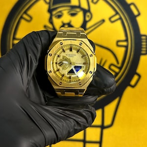 May include: A gold-toned Casio G-Shock watch featuring an octagonal bezel with eight visible screws, a metallic gold dial with analog hands, and a digital display window. The watch has a brushed metal finish and a matching gold-toned metal link band.