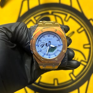 May include: A gold-toned metal wristwatch featuring an octagonal bezel with eight visible screws. The watch face is light blue with analog hands, a digital display window showing 07, and the text G-SHOCK CASIO printed on the dial surface.