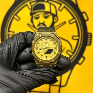 May include: A gold-toned G-Shock Casio wristwatch with an octagonal bezel, featuring a yellow analog-digital dial with luminous hands, a digital display window, and a black strap. The watch face displays the text G-SHOCK CASIO.