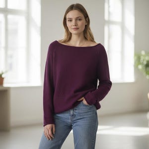 Könnte beinhalten: Ein tiefvioletter Pullover mit weitem Ausschnitt und langen Ärmeln. Der Pullover wird mit einer blauen Jeans kombiniert. Das Model steht in einem hellen Raum mit großen Fenstern und einer Topfpflanze im Hintergrund.