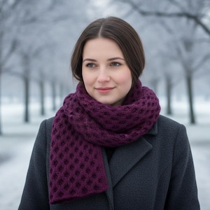May include: A woman wearing a dark purple knitted scarf and a dark gray coat. The scarf has a textured pattern. The background shows a snowy path and trees, suggesting a cold winter day. The scarf appears to be handmade.