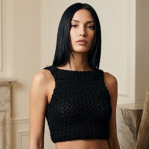 Peut inclure: Un haut court noir sans manches en crochet. Le haut a une encolure carrée et un aspect texturé. Le modèle de l'image a de longs cheveux noirs raides et regarde directement l'appareil photo. L'arrière-plan comprend une cheminée et des rideaux à motifs.