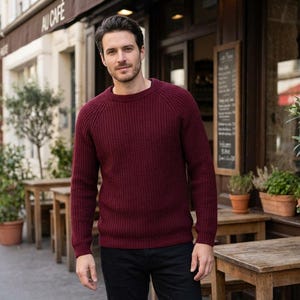May include: A burgundy ribbed knit sweater with a crew neck. The sweater is worn by a person standing outside a cafe. The cafe has wooden tables and chairs. The sign above the cafe says "AU CAFE".