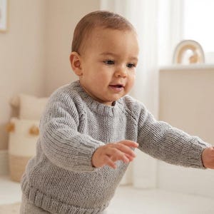 May include: A baby wearing a light gray knit sweater and matching pants. The baby is reaching out with both hands. The sweater has a crew neck and ribbed cuffs. The baby is in a room with neutral tones.