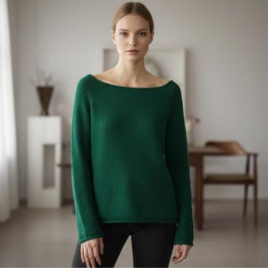 Könnte beinhalten: Eine Frau trägt einen langärmeligen, smaragdgrünen Pullover mit weitem U-Boot-Ausschnitt. Der Pullover wird mit einer schwarzen Hose kombiniert. Der Hintergrund umfasst einen weißen Vorhang, einen weißen Sockel und einen Holztisch.