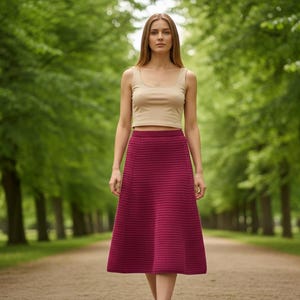 Op de afbeelding: Een vrouw draagt een beige crop top en een bordeauxrode midi-rok, lopend op een pad omzoomd door groene bomen. De rok heeft een getextureerde afwerking. De stijl is casual en elegant.