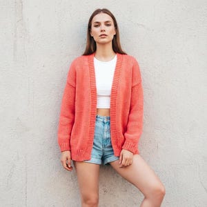 May include: A vibrant coral cardigan with long sleeves, paired with a white crop top and denim shorts. The outfit is set against a neutral wall. The cardigan's colour provides a striking contrast. The overall style suggests a casual, everyday aesthetic.
