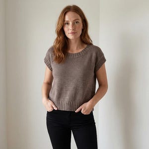 May include: A brown knitted top with short sleeves and a ribbed texture. The top features a crew neck and a slightly cropped design. The model is wearing black trousers. The background is a plain white wall.