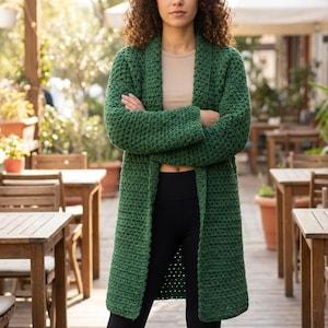 May include: A long, emerald green, open-front cardigan. The chunky knit sweater has a shawl collar and long sleeves. The person is wearing a beige top and black leggings. The background includes outdoor seating and greenery.