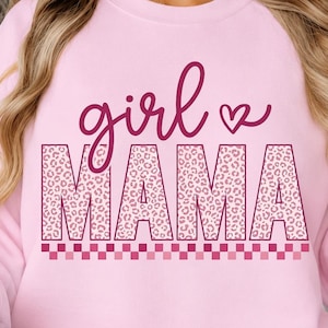 May include: A light pink crewneck sweatshirt featuring the text girl MAMA in a dark pink script and block font. The block letters have a pink leopard print pattern, and a small heart icon is positioned next to the word girl above the text.