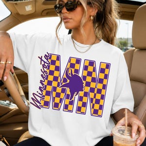 May include: A white short-sleeved t-shirt featuring a large graphic with the text Minnesota and MN in a purple and gold checkered pattern. The design includes a purple Viking helmet silhouette integrated into the letters on the front of the shirt.