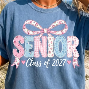 Könnte beinhalten: Ein blaues kurzärmeliges T-Shirt mit einem großen Grafikdruck einer rosa-blauen, blumig gemusterten Schleife über dem Wort SENIOR im Patchwork-Stil, mit dem Text Class of 2027 und kleinen rosa Herzen unter dem Hauptschriftzug.
