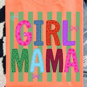 May include: An orange t-shirt featuring a green vertical striped background and the text GIRL MAMA in large, multi-colored block letters with various patterns including polka dots, stars, and wavy lines, accented by small light blue and pink bow icons.