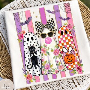 Peut inclure: Un t-shirt blanc avec un motif de trois fantômes sur des rayures verticales roses et violettes. Les fantômes sont ornés de pois noirs, de motifs floraux et de carreaux orange, agrémentés de chauves-souris, d'un chat noir et d'une citrouille.