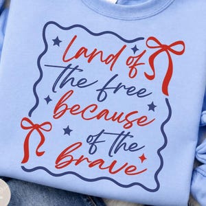 Puede incluir: Sudadera azul claro con la frase "Land of the free because of the brave" en rojo y azul. El diseño incluye lazos rojos y estrellas dentro de un borde azul ondulado. La sudadera está hecha de un material suave.