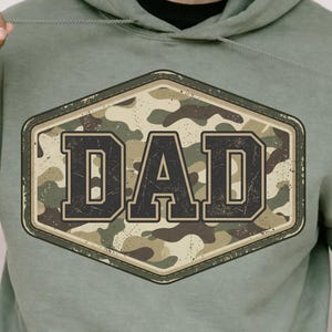 May include: Sage green hoodie with a large, distressed, camouflage-patterned patch that reads "DAD" in bold, dark letters. The patch is framed by a dark border. The hoodie has a drawstring and a front pocket.